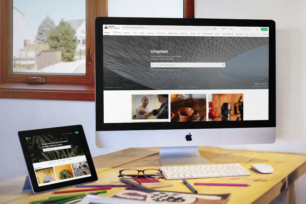 A Mac computer and a tablet sit on a rustic desk displaying the homepage of Unsplash stock photo site. 